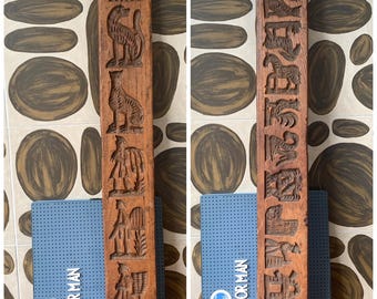 May include: Two antique wooden molds with carved relief designs. One features figures and animals, the other has text and symbols. The molds are brown and rest on a blue book with the text "DE KLEINE NOOR MAN". The background has a brown and white pattern.