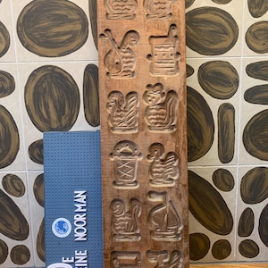 May include: A wooden cookie mould with eight different designs carved into it. The designs include a rooster, a boat, a bucket, and a fish. The mould is brown and has a worn look.
