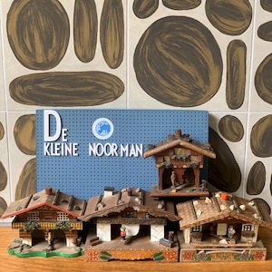 May include: Three wooden chalet-style miniature houses with brown roofs and painted details, positioned in front of a blue pegboard sign that reads DE KLEINE NOORMAN. Each house features small figurines, balconies, and floral accents on the base.