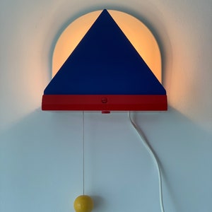 Puede incluir: Una lámpara de pared triangular azul y roja con un semicírculo blanco detrás. La lámpara tiene una cadena de tracción amarilla.