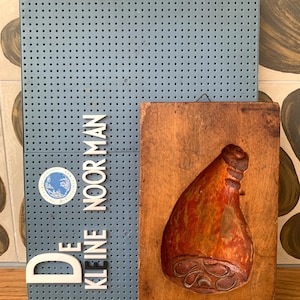 May include: A vintage wooden meat mold with a carved ham design, next to a blue pegboard with the text "DE KLEINE NOORMAN" in white letters. The mold is brown with red and orange hues.