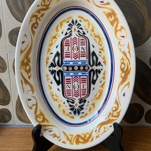 May include: Oval ceramic serving platter with a decorative design. The white platter features a central emblem with red, blue, and black details, framed by a blue and gold border. The rim has a gold scrollwork pattern.