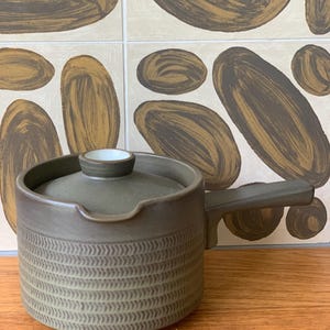 May include: A dark olive green ceramic teapot with a matching lid and a single side handle. The lower portion of the cylindrical body features a textured, repeating herringbone pattern, while the upper rim includes a small spout notch.
