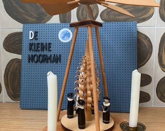 May include: A wooden Christmas pyramid with a rotating propeller, a spiral-shaped tree, and six figures standing around the base. Two white candles are in candle holders on the bottom level. The pyramid is on a wooden surface. The text "DE KLEINE NOORMAN" is visible on a blue background.