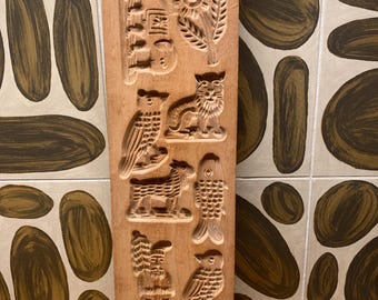 May include: A rectangular, wooden cookie mold with carved designs. The mold features various images, including a house, flower, owl, cat, sheep, fish, Santa Claus, and a bird. The wood has a natural, light brown color.