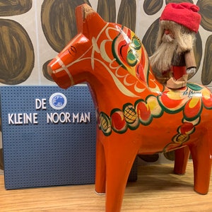 May include: A traditional Swedish Dala horse, painted in red, yellow, green and white, with a small gnome riding on top. The horse is standing on a wooden surface. The text "DE KLEINE NOOR MAN" is visible on a blue sign behind the horse.