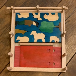 Op de afbeelding: Houten puzzel met een blauwe en groene landschapsscène en een rode houten basis. De puzzelstukken zijn gevormd als dieren en wolken.