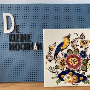 May include: A blue pegboard with the words "De kleine Noorman" spelled out in white and black letters. Next to the pegboard is a white ceramic tile with a yellow and blue bird surrounded by brown, yellow, and blue flowers.