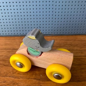 May include: A wooden toy car featuring a natural light-colored wood body and four bright yellow wheels with silver centers. A gray elephant figure with a green shirt and yellow bowtie is seated in the center of the car body, facing forward.