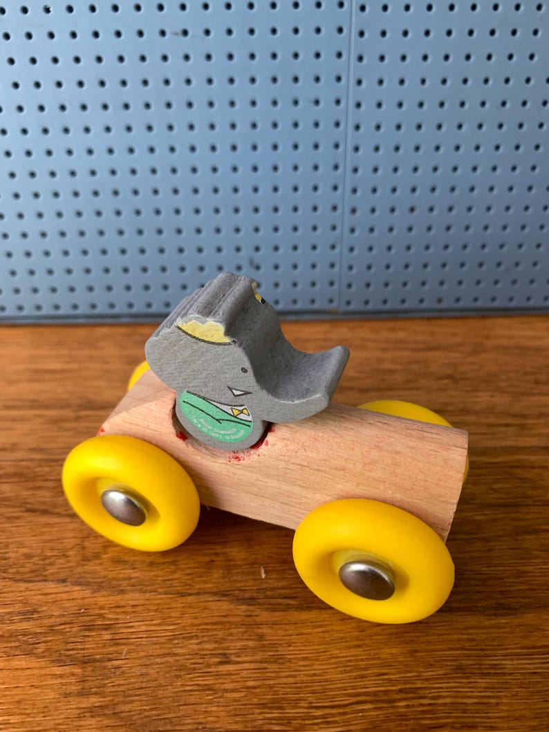 May include: A wooden toy car featuring a natural light-colored wood body and four bright yellow wheels with silver centers. A gray elephant figure with a green shirt and yellow bowtie is seated in the center of the car body, facing forward.
