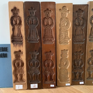 May include: Six wooden cookie molds with different designs of people in traditional clothing. The molds are numbered 1 through 6. The text "DE KIEINE NOORKMAN" is visible in the background.