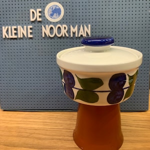 May include: A ceramic bowl with a lid, featuring a blue and green floral pattern, sits atop a brown pedestal. The bowl has a white base and a blue lid. The background includes the text "DE KLEINE NOORMAN".