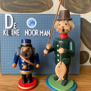 May include: Two wooden figurines: a fisherman with a fishing rod and a fish, and a soldier. The fisherman wears a green coat and brown hat. The soldier is in a blue uniform. The background features the text "De Kleine Noorman".