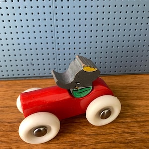 May include: A toy car featuring a bright red wooden body and four white plastic wheels with silver metal centers. A gray elephant figure with a yellow ear and green collar is attached to the top of the car, serving as the driver of the vehicle.