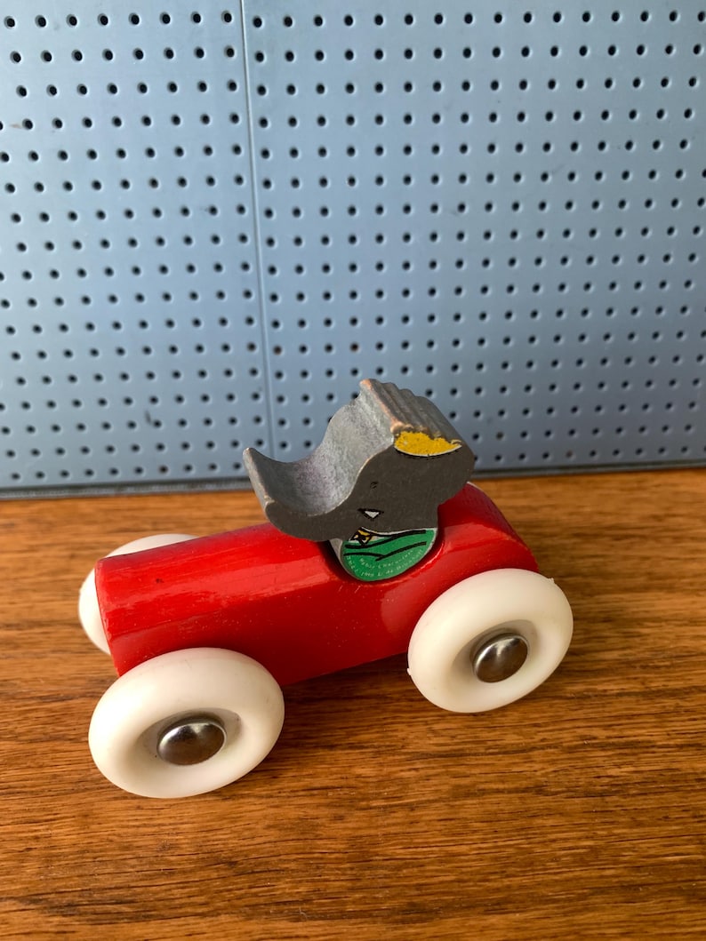 May include: A toy car featuring a bright red wooden body and four white plastic wheels with silver metal centers. A gray elephant figure with a yellow ear and green collar is attached to the top of the car, serving as the driver of the vehicle.