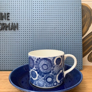 May include: A white ceramic mug with a blue circular pattern sits on a matching blue saucer. The mug has a white handle. The background includes a blue pegboard with the text "DE KLEINE NOORMAN".