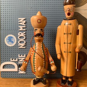 May include: Two wooden figurines. One is a man with a mustache, wearing a hat and a decorated tunic. The other is a man in a long coat, holding a stick and a small barrel. The background is a blue pegboard with the text "DE KLEINE NOORMAN".