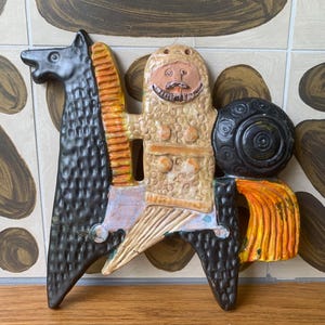 May include: A ceramic wall plaque depicting a knight in textured tan armour riding a black horse. The knight holds a round black shield and has a painted face with a moustache. The horse features a yellow mane and tail, with orange accents.