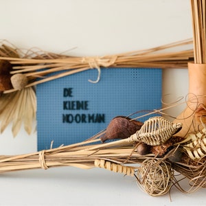 May include: A blue pegboard with the text "DE KLEINE NOORMAN" in black lettering.  Dried natural grasses and other plant materials are arranged in front of the board.