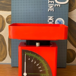 May include: A bright orange mechanical kitchen scale featuring a rectangular weighing tray on top and a curved display panel. The display shows a green scale with white numbers ranging from 50 to 300, a white needle, and the EKS brand logo at the bottom.