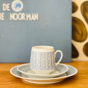 May include: A white ceramic coffee cup with a gray and white geometric pattern. The cup is sitting on a white saucer with a gray and white geometric pattern. The saucer is sitting on a wooden table.