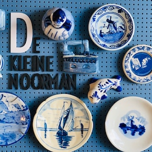 May include: A blue and white Delftware collection featuring a variety of decorative items, including a small clock, a figurine of a woman, a small iron, a figurine of a fish, and several plates with blue and white designs. The items are displayed on a blue pegboard with the text "DE KLEINE NOORMAN" in black letters.