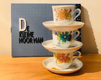 Peut inclure: Trois tasses à thé et soucoupes vintage empilées avec des motifs floraux orange, jaune et bleu. Les tasses sont blanches avec une anse incurvée. Un panneau perforé bleu avec le texte "De Kleine Noorman" est en arrière-plan.