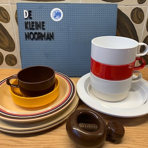 May include: A set of four white ceramic cups stacked with a red band, a brown ceramic cup with a yellow saucer, a set of four beige and brown striped saucers, and a brown nutcracker. The cups and saucers are on a wooden surface. A blue sign with white lettering reads "DE KLEINE NOORMAN".