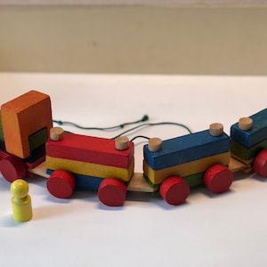 May include: A colorful wooden toy train with a green, orange, red, yellow, blue, and green engine and cars. The train has red wheels and a yellow wooden person.