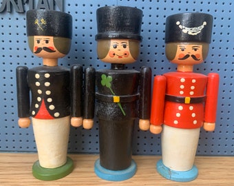 Könnte beinhalten: Drei Holzfiguren von Menschen, die verschiedene Hüte und Uniformen tragen. Die Figur links trägt einen schwarzen Hut mit einem goldenen Stern, eine schwarze Jacke mit weißen Punkten und eine weiße Hose. Die Figur in der Mitte trägt einen schwarzen Zylinder, eine schwarze Jacke mit einem grünen Kleeblatt und eine schwarze Hose. Die Figur rechts trägt einen schwarzen Hut mit einem weißen Band, eine rote Jacke mit weißen Punkten und eine weiße Hose.