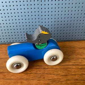 May include: A wooden toy car painted bright blue with four white wheels featuring silver center caps. A gray elephant figure with a yellow ear and a green shirt is attached to the top of the car body. The toy has a smooth finish and simple design.