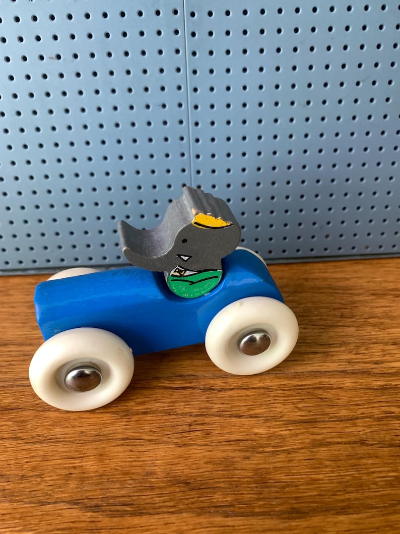 May include: A wooden toy car painted bright blue with four white wheels featuring silver center caps. A gray elephant figure with a yellow ear and a green shirt is attached to the top of the car body. The toy has a smooth finish and simple design.