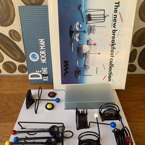 Puede incluir: Un conjunto de piezas de servicio de desayuno de metal negro con acentos esféricos coloridos. El juego incluye cuchilleros, hueveras y otros accesorios para servir. La caja dice "The new breakfast collection."