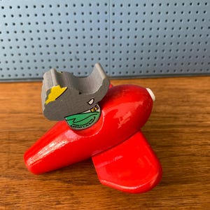 May include: A bright red wooden toy airplane featuring a gray elephant character figure seated in the cockpit. The elephant wears a green shirt and a yellow hat. The text on the figure reads "BARBAR CHARACTERS (C) 1995 L. de Brunhoff".