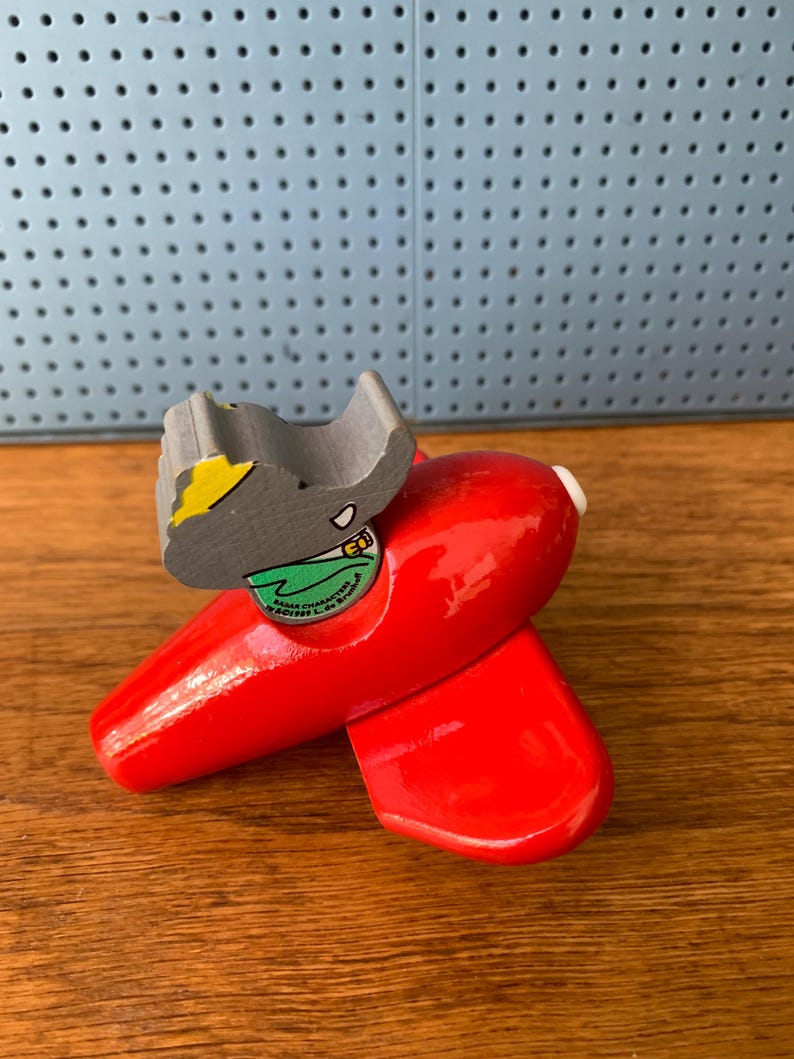 May include: A bright red wooden toy airplane featuring a gray elephant character figure seated in the cockpit. The elephant wears a green shirt and a yellow hat. The text on the figure reads "BARBAR CHARACTERS (C) 1995 L. de Brunhoff".