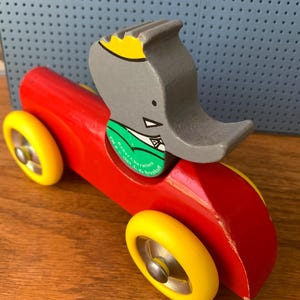 May include: A wooden toy car painted bright red with four yellow wheels and a silver metal hubcap on each. A gray elephant figure with a yellow crown and a green collar is attached to the top. The green collar features the text Babar Characters TM & &copy; 1989 L. de Brunhoff.