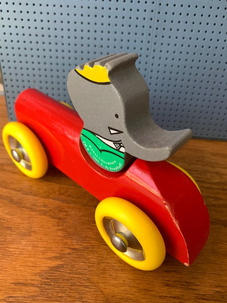 May include: A wooden toy car painted bright red with four yellow wheels and a silver metal hubcap on each. A gray elephant figure with a yellow crown and a green collar is attached to the top. The green collar features the text Babar Characters TM & &copy; 1989 L. de Brunhoff.