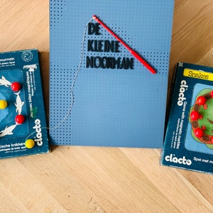 May include: Two blue and yellow magnetic game boards with the text "clacto" and "Snakes" on them. The game boards are on a wooden surface. A blue board with the text "DE KLEINE NOORMAN" is in the background.