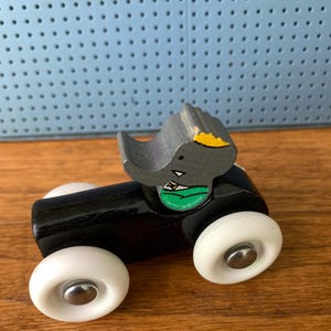 May include: A small wooden toy vehicle with a black cylindrical body and four white plastic wheels with silver centers. A gray wooden elephant figure with a yellow ear and a green circular sticker featuring the text Laurent de Brunhoff sits on top.