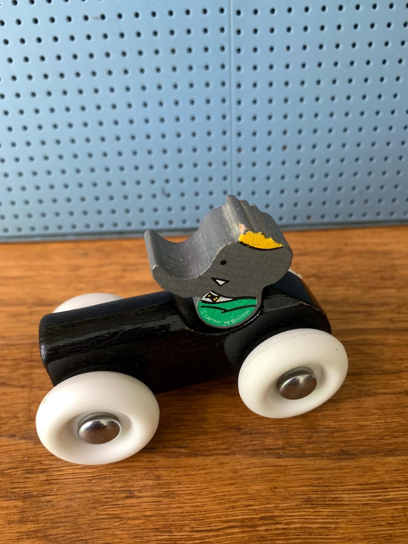 May include: A small wooden toy vehicle with a black cylindrical body and four white plastic wheels with silver centers. A gray wooden elephant figure with a yellow ear and a green circular sticker featuring the text Laurent de Brunhoff sits on top.