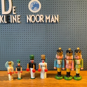 Peut inclure: Une collection de figurines en bois, dont des casse-noisettes et des anges, est exposée sur une surface en bois. Les figurines sont peintes de différentes couleurs, dont le bleu, le vert, le rose et l'or. Le texte "De Kleine Noor Man" est visible en arrière-plan.
