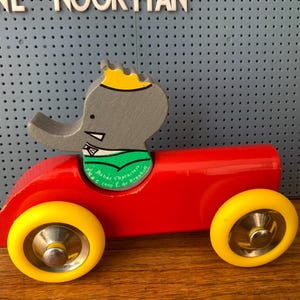 May include: A wooden toy car painted bright red with two yellow wheels and a silver hubcap on each. A gray elephant figure with a yellow crown and green scarf is seated in the center. The green scarf features the text Babar Characters 1999 L de Brunhoff.