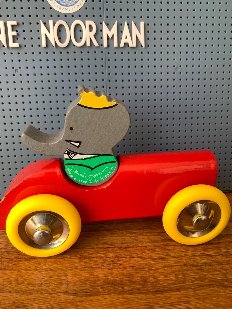 May include: A wooden toy car painted bright red with two yellow wheels and a silver hubcap on each. A gray elephant figure with a yellow crown and green scarf is seated in the center. The green scarf features the text Babar Characters 1999 L de Brunhoff.