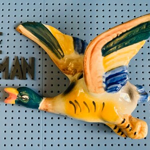 May include: A ceramic duck figurine in flight, with wings spread wide. The duck features a yellow body, blue, green, and pink accents on the wings, and a green head. The duck is mounted on a blue pegboard.