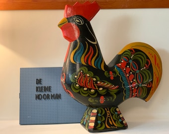May include: A black wooden rooster with colorful painted details. The rooster has a red comb and wattle, yellow tail feathers, and blue, green, and red painted designs on its body. The rooster is standing on a white surface with a blue and white sign that says "DE KLEINE NOOR MAN".