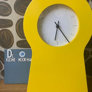 May include: A bright yellow keyhole-shaped clock with black hands and a white face. The clock face has black numbers 1 through 12.