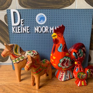 May include: Two wooden horse figurines with painted floral patterns and two rooster figurines with vibrant red, orange, and blue painted details. The items are positioned in front of a blue pegboard with the white text DE KLEINE NOORMAN.