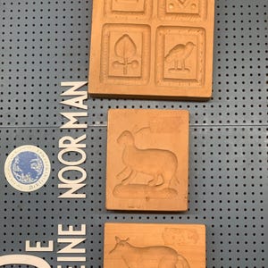 May include: Three wooden cookie cutters with different designs. The top cutter has four squares with a lamb, a diamond pattern, a tree, and a bird. The middle cutter has a rabbit. The bottom cutter has a fox. The cutters are displayed on a grey pegboard with the text "De Kleine Noorman" in white letters.