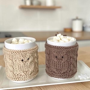 Könnte beinhalten: Zwei weiße Keramikbecher gefüllt mit Marshmallows, jeweils umhüllt von einer gestrickten Eulen-Design-Hülle. Eine Hülle ist beige, die andere braun. Die Becher stehen auf einem weißen Tablett.