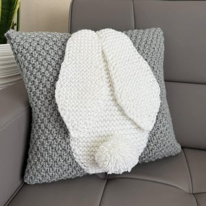 May include: A white knitted bunny rabbit pillow with a gray knitted pillow behind it. The bunny pillow has a pom-pom tail.